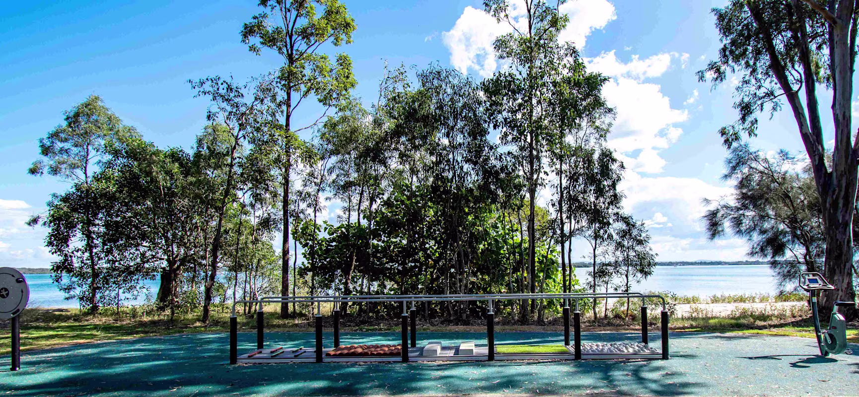 Outdoor gym with rails and exercise equipment on blue rubber surface, shaded by tall trees beside a calm lake under blue sky