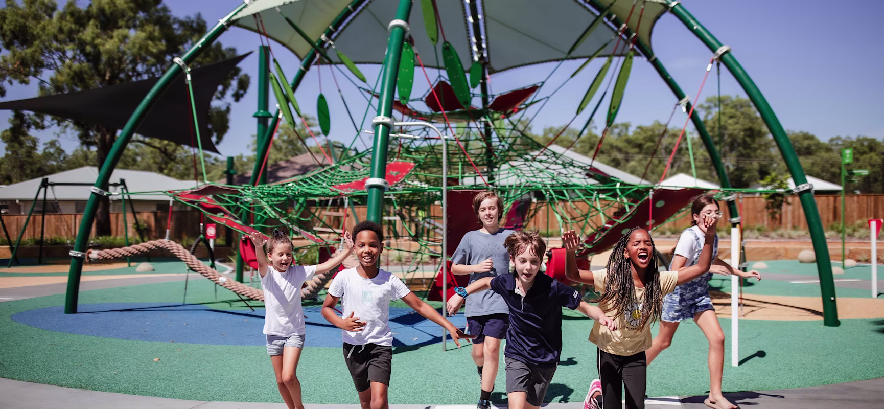 Children run toward the camera at a colorful outdoor playground with a large green climbing structure and shade sails.