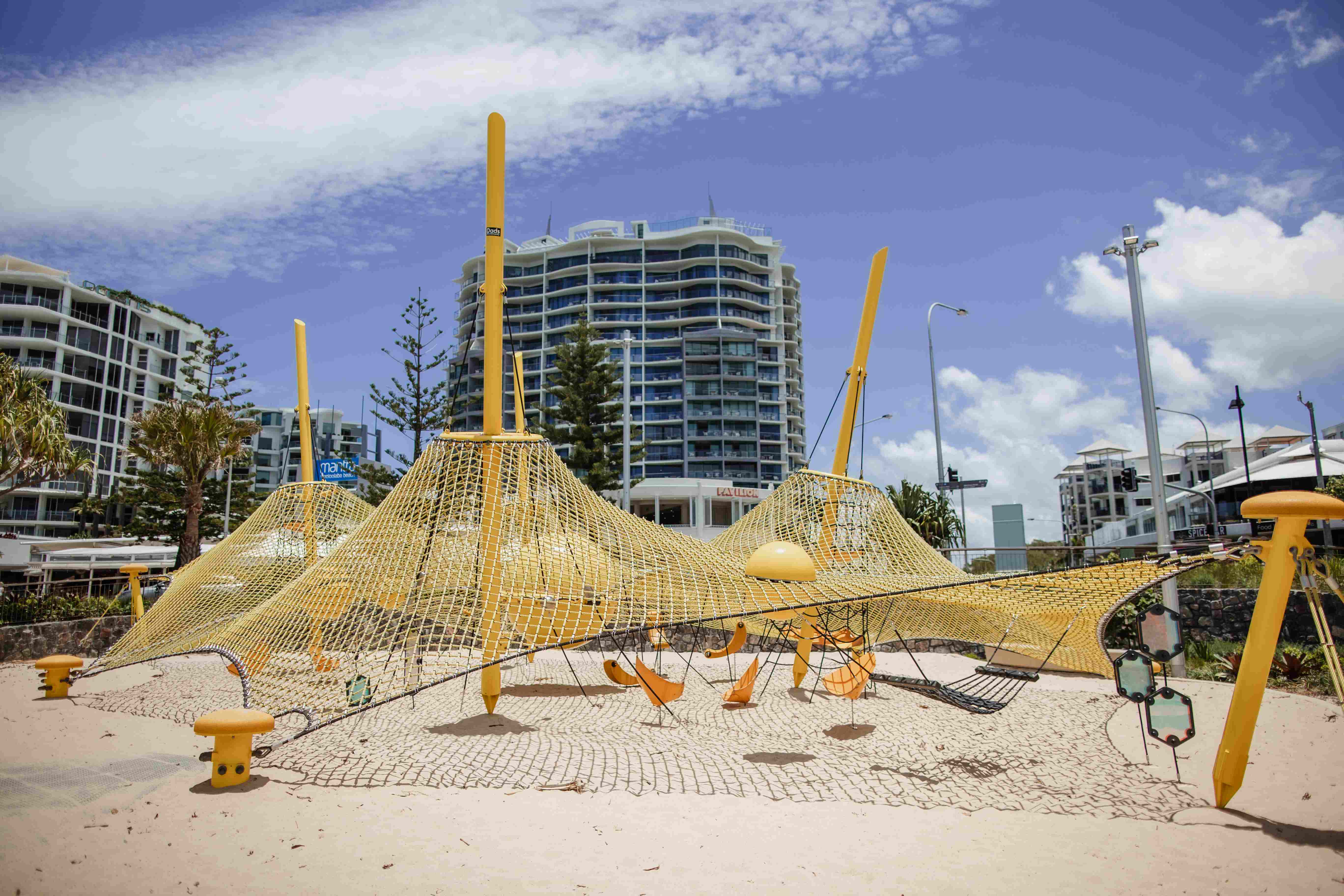 Yellow rope climbing playground on sandy ground, with high-rise apartments, palm trees and a blue sky with clouds in the background.