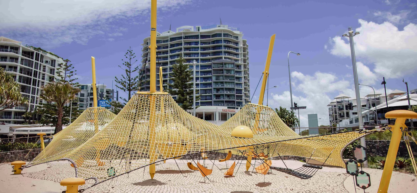 Yellow rope climbing playground on sandy ground, with high-rise apartments, palm trees and a blue sky with clouds in the background.