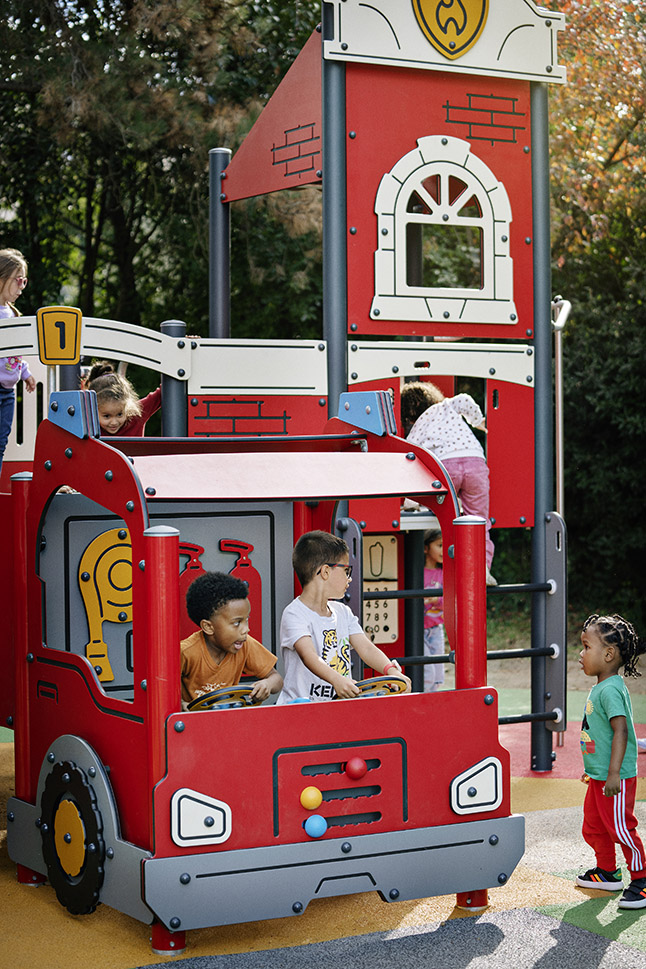 Children play on a red fire truck and station themed playground structure outdoors, surrounded by trees and colourful safety flooring