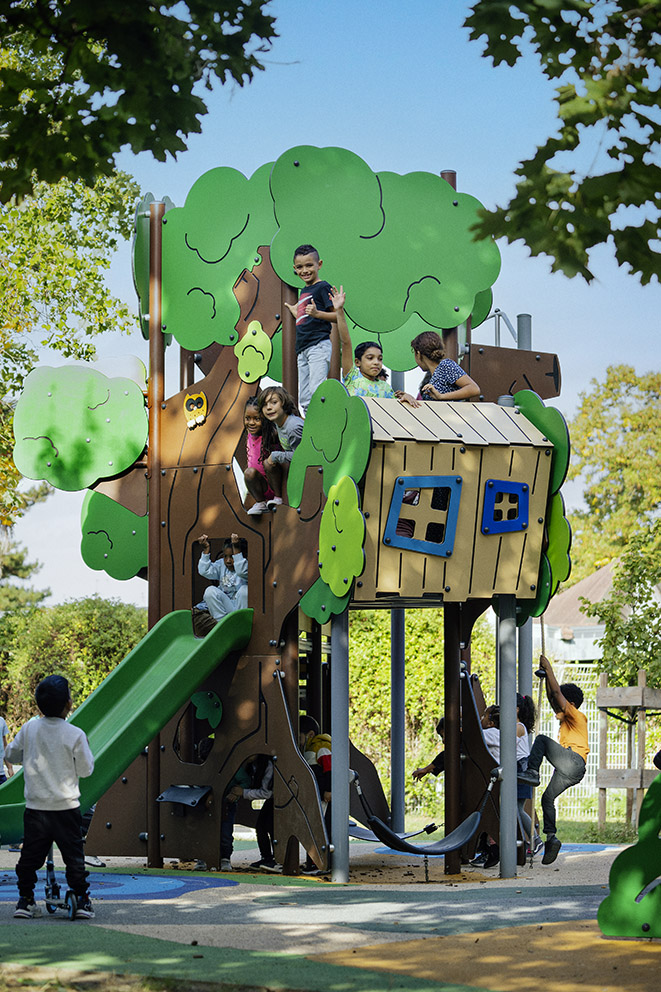 Children play on a red fire truck and station themed playground structure outdoors, surrounded by trees and colourful safety flooring