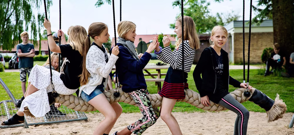 Children sit on a large rope swing in a playground, facing each other and talking, with trees and other kids in the background