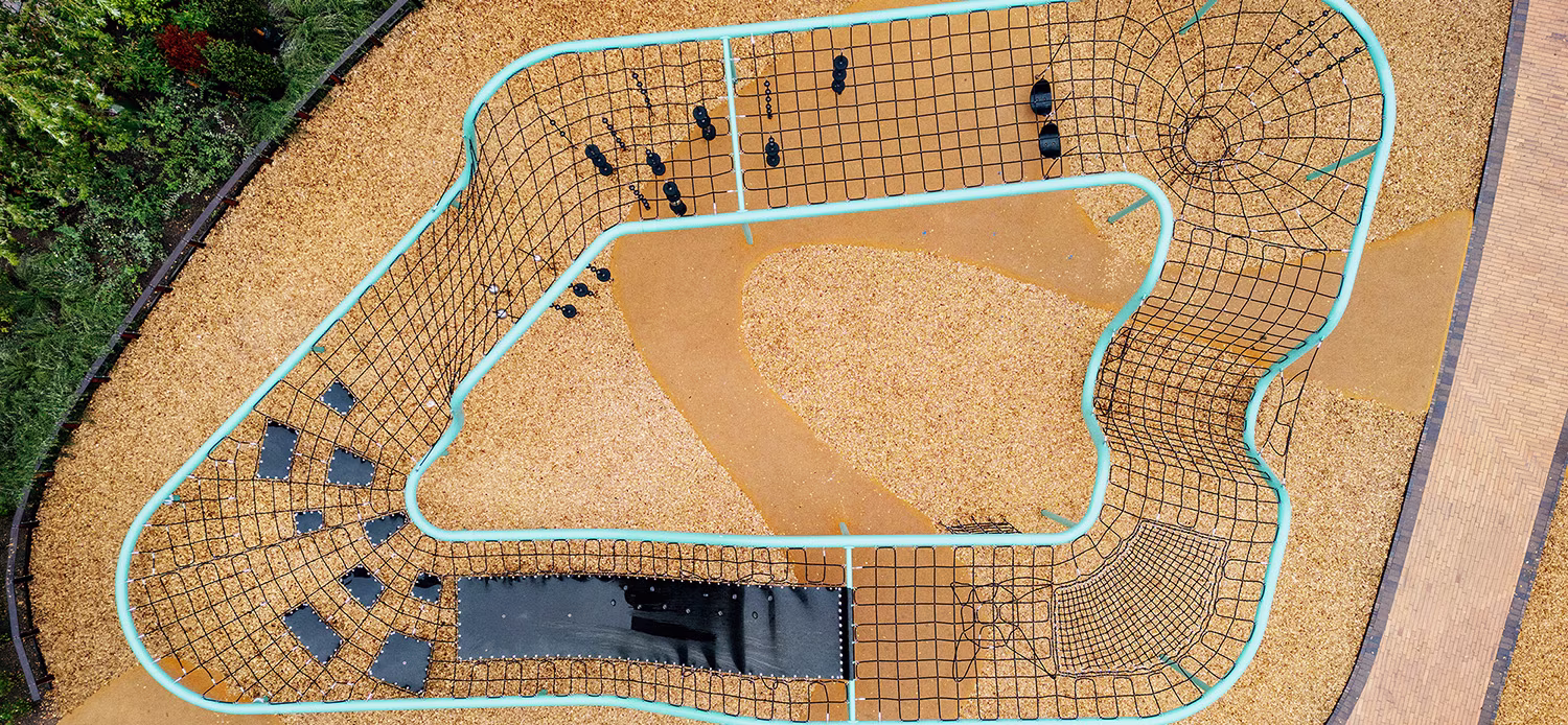 Aerial view of a modern playground with turquoise climbing nets, rope tunnel and yellow rubber surface bordered by plants and paths