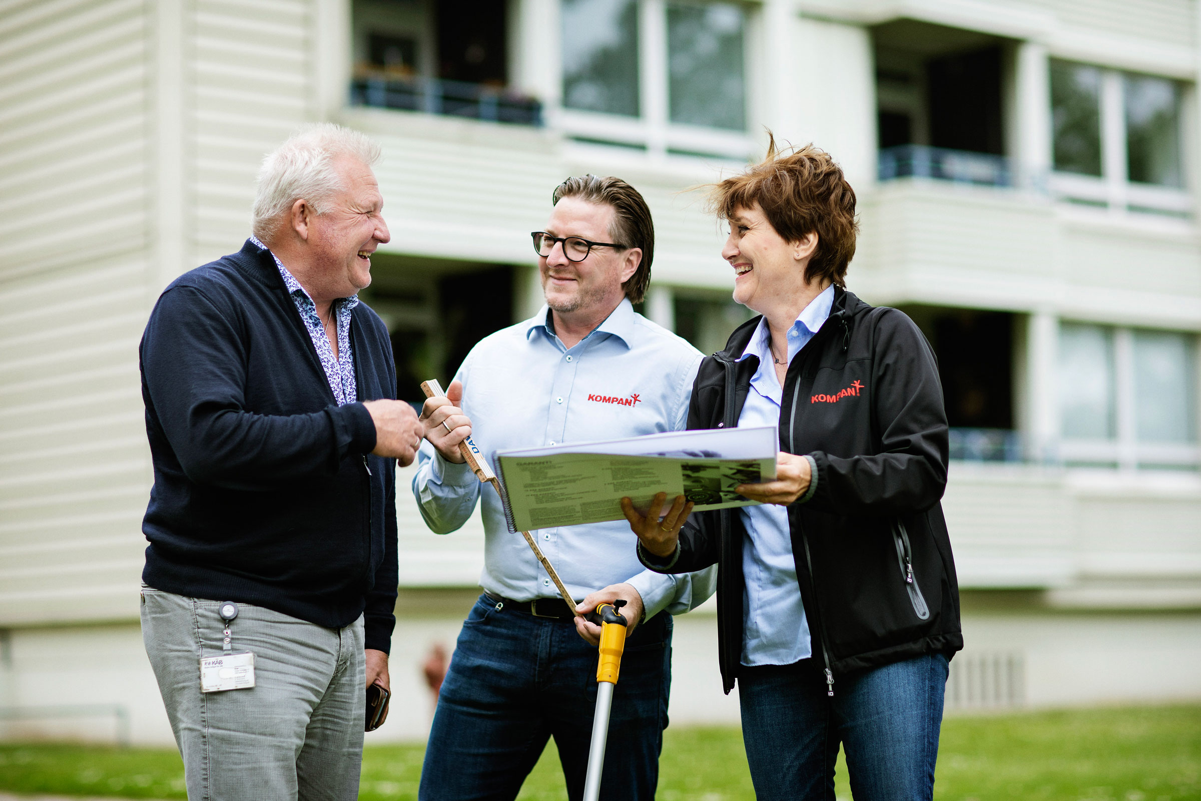 Three people stand outside an apartment building, discussing plans while holding documents and a measuring tool on a grassy area