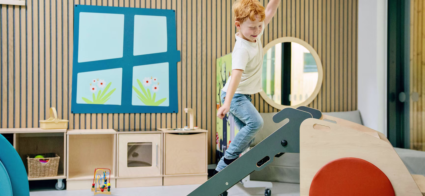 Child with blurred face climbing indoor play ramp in bright, modern playroom with wooden walls and colorful decor