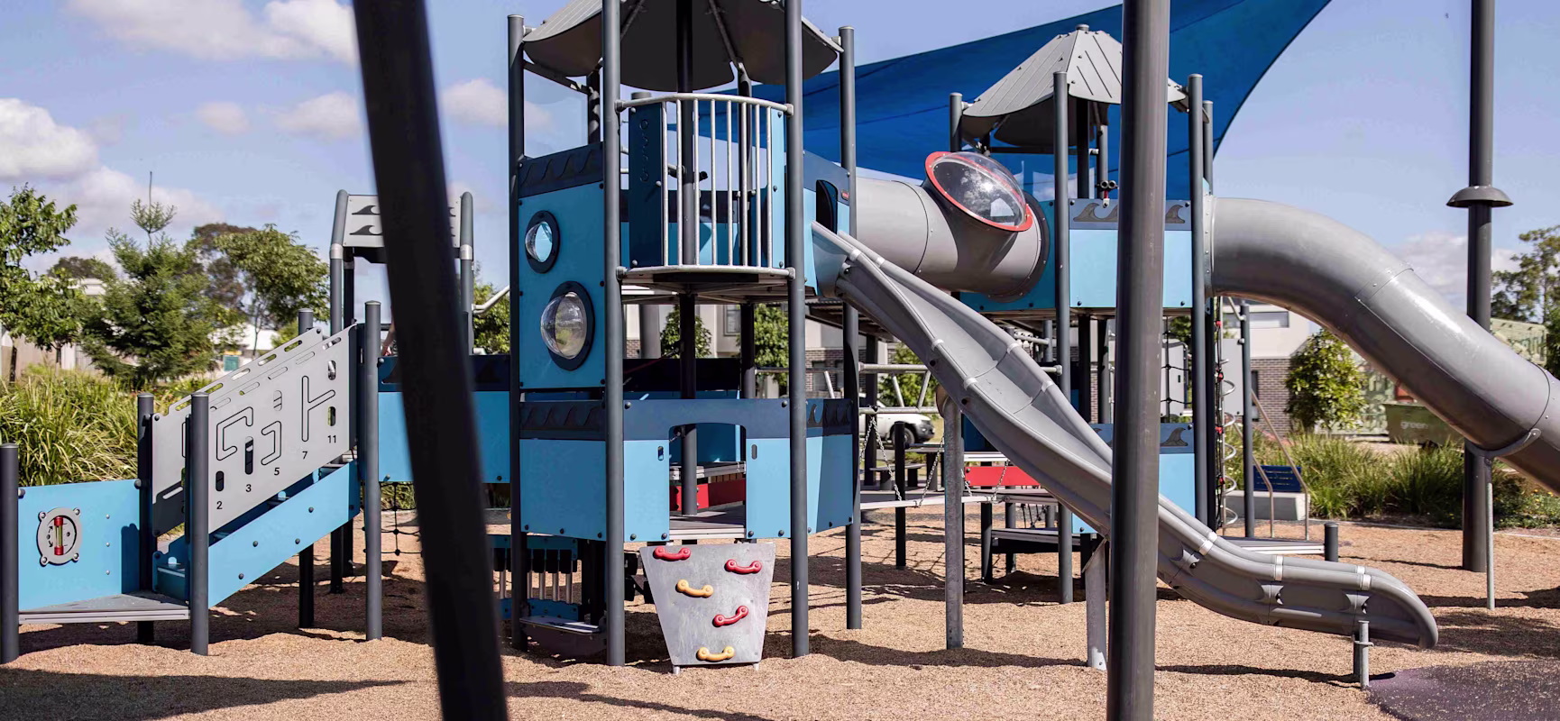 Modern blue playground with towers, tube tunnels, and long grey slides on mulch under a sunny sky and shade sail.