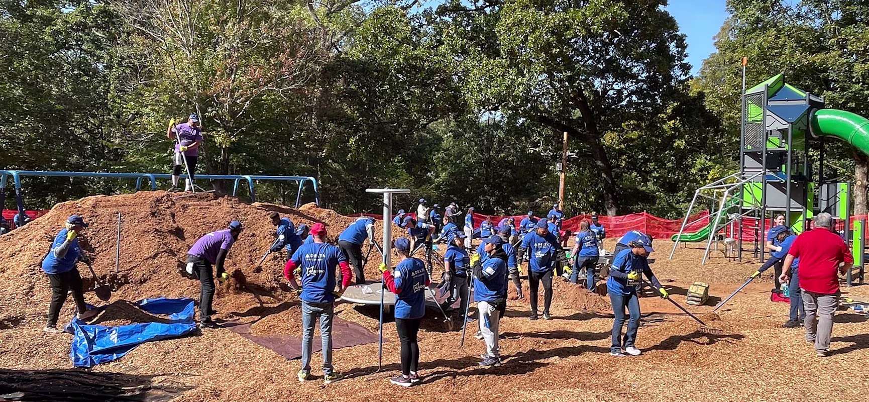 Volunteers in blue shirts spread wood chips at a playground construction site surrounded by trees and slides on a sunny day