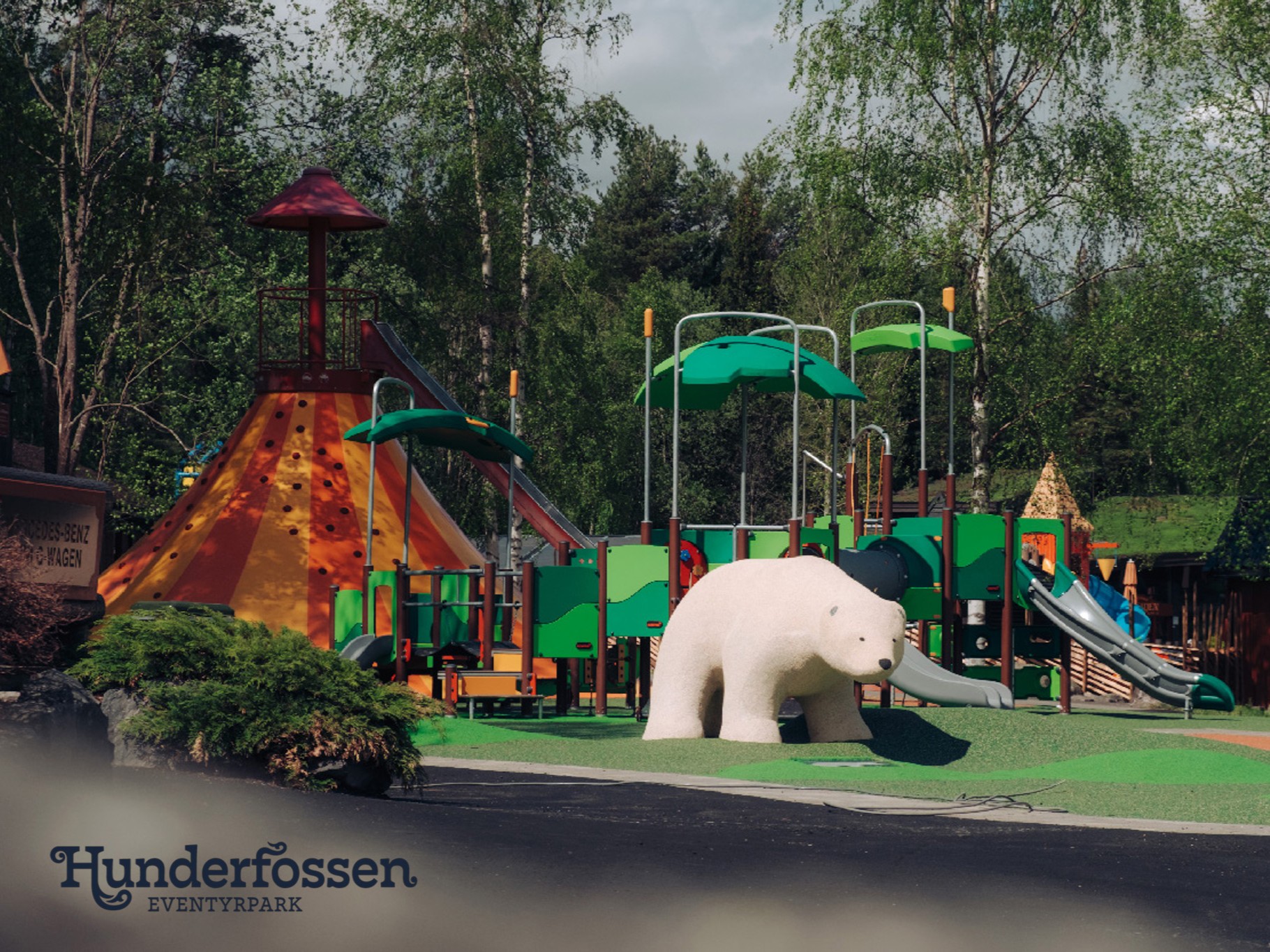 Colorful playground with slides and climbing structures, a large white polar bear sculpture, and green trees in the background.