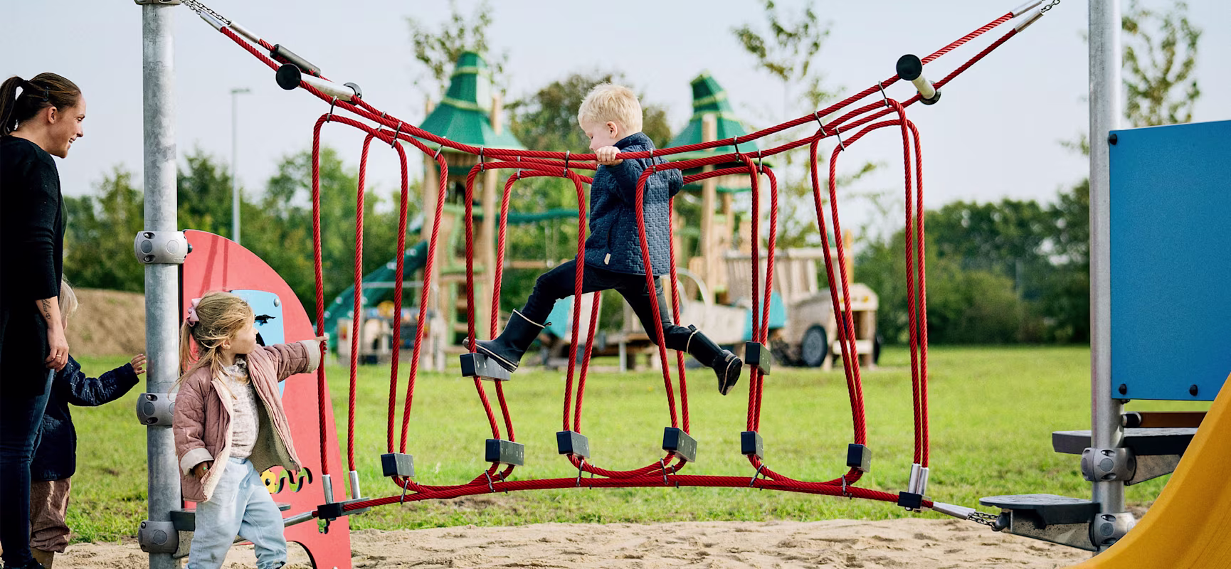 Children playing on a red rope bridge at a playground, with an adult nearby and slides and climbing structures in the background