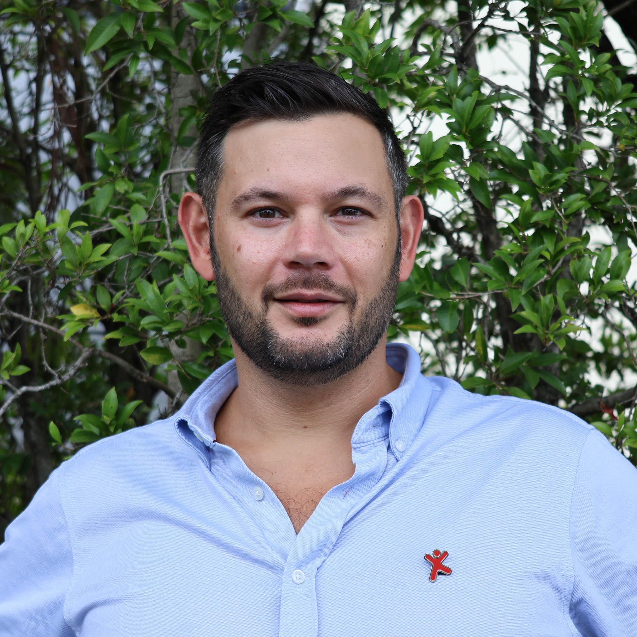 Person with blurred face in light blue button-up shirt, standing in front of leafy green bushes, small red logo on chest.