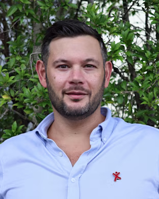 Person with blurred face in light blue button-up shirt, standing in front of leafy green bushes, small red logo on chest.