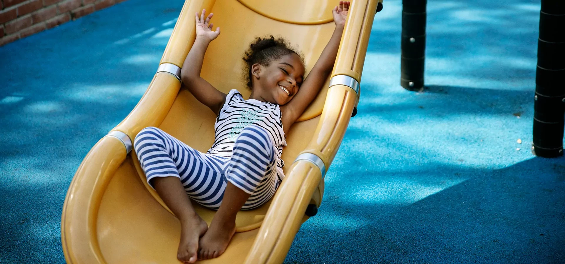 A girl in a striped outfit sliding down a yellow slide with bright blue surfacing underneath. Including bright colours within your playground construction details can produce vibrant and enticing results.