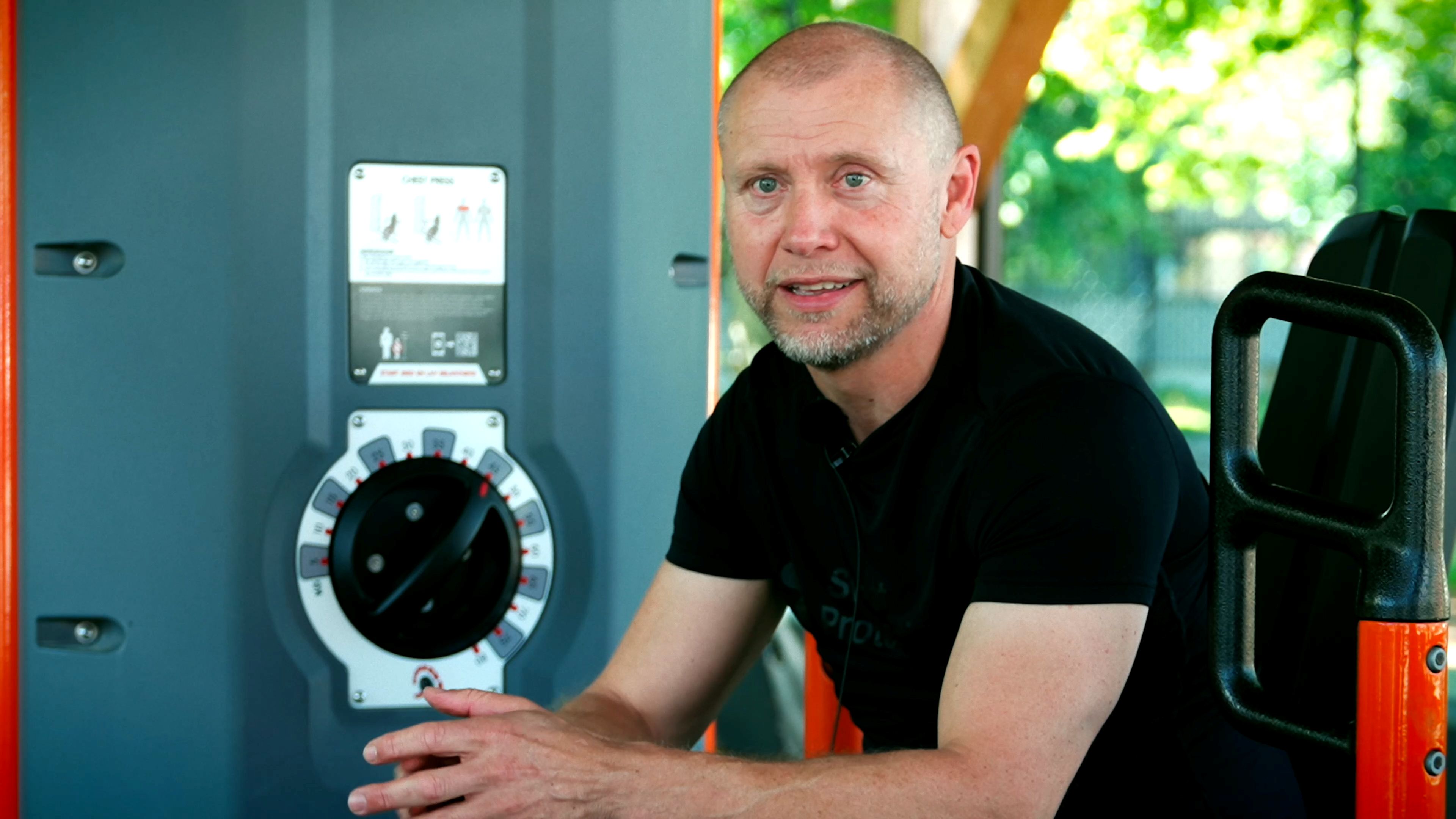 Person in black shirt sitting on outdoor gym machine, hands clasped, next to grey control panel with dial and exercise diagram