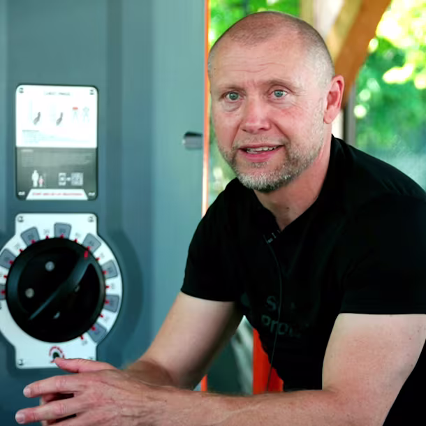 Person in black shirt sitting on outdoor gym machine, hands clasped, next to grey control panel with dial and exercise diagram