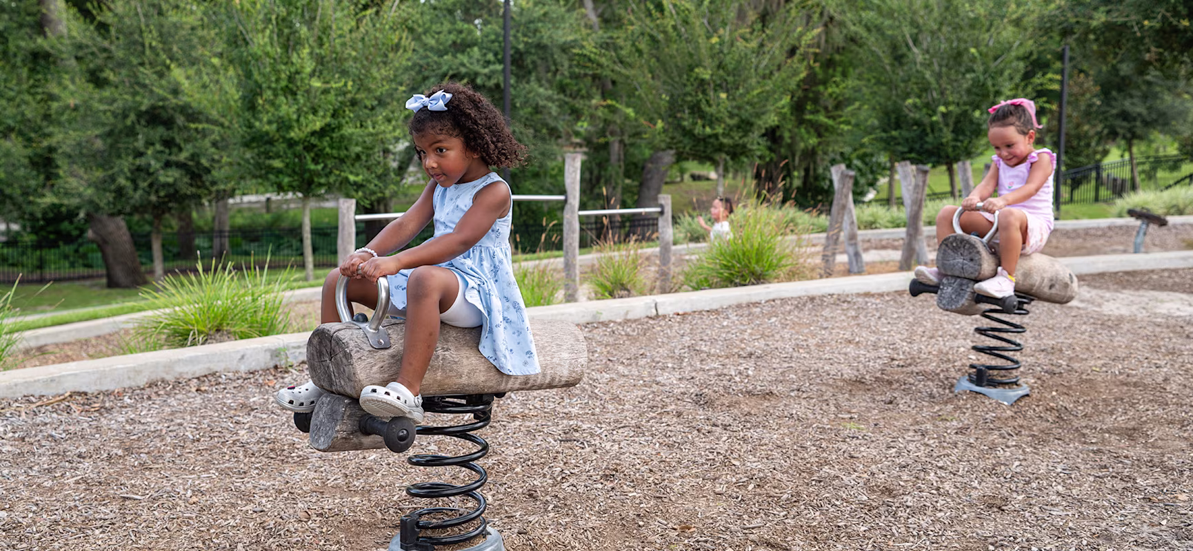 Two young girls ride separate spring rockers on a playground, surrounded by wood chips and green trees in the background.