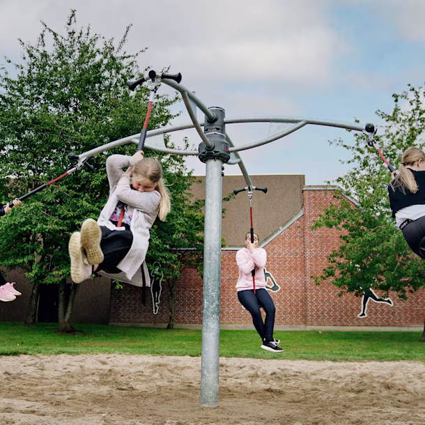 4 tween girls swinging together on a carousel. Each has their own seat. The surfacing underneath is safety sand. Did you know the type of surfacing you choose for your playground construction project can change the aesthetic of the final product?