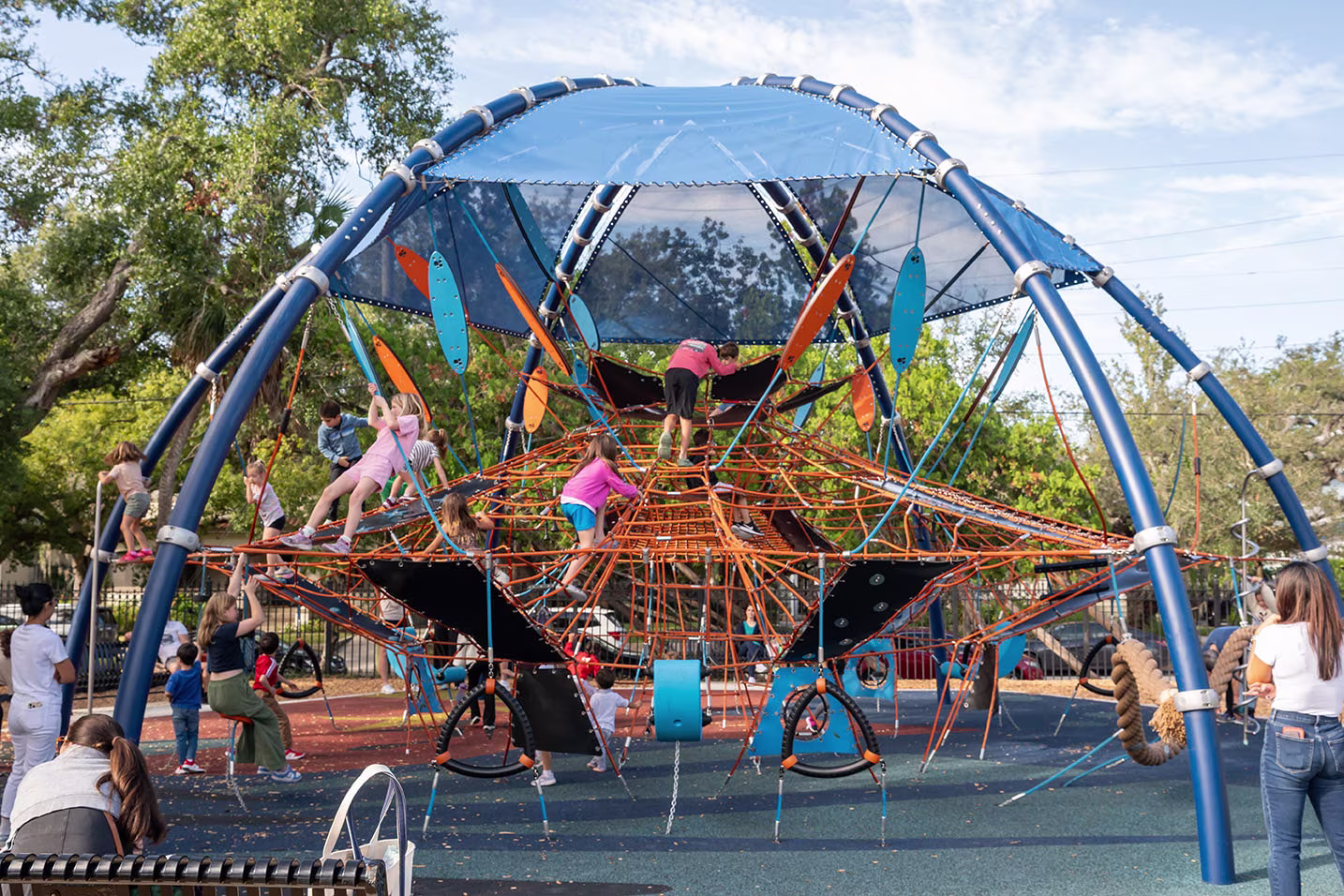 Children climb a large blue and orange rope playground structure while adults watch under a bright sky and surrounding trees