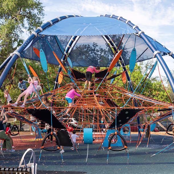 Children climb a large blue and orange rope playground structure while adults watch under a bright sky and surrounding trees