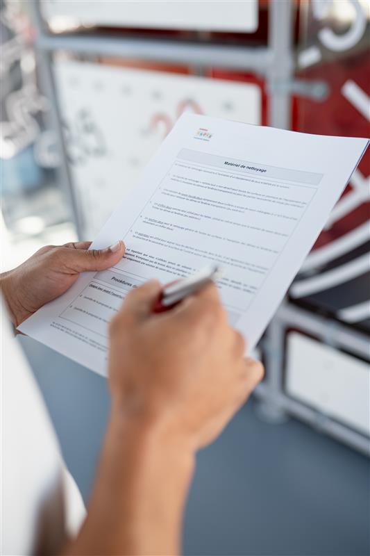 Person holding and filling out a printed form with a pen, viewed from above, with blurred office equipment in the background