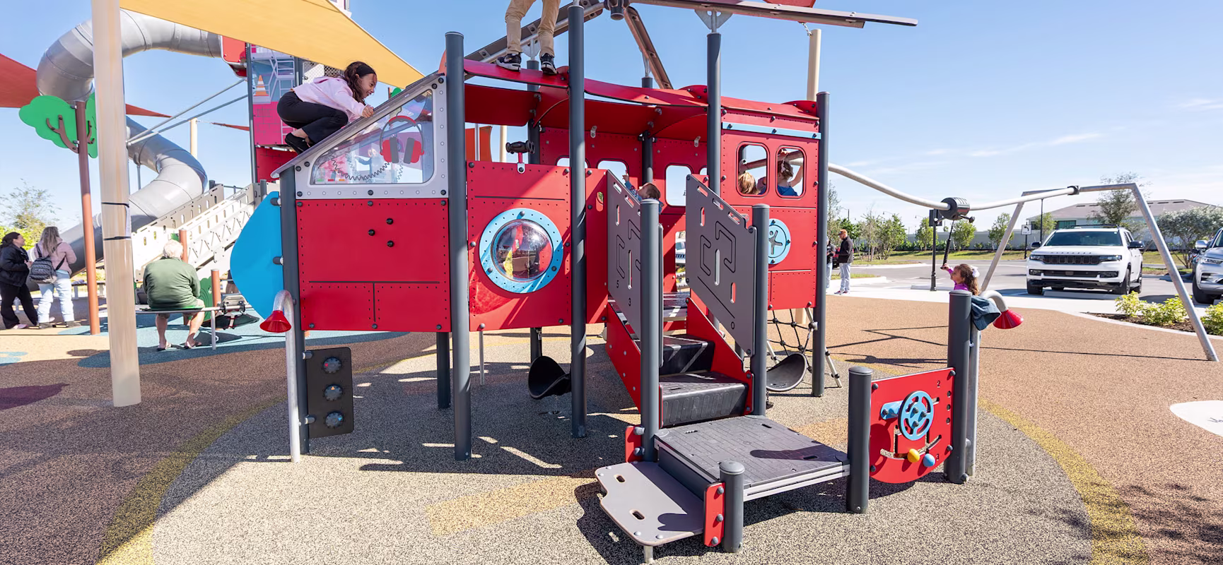 Children climbing a bright red airplane-themed playground structure under shade sails on a sunny day, with adults and cars in the background.
