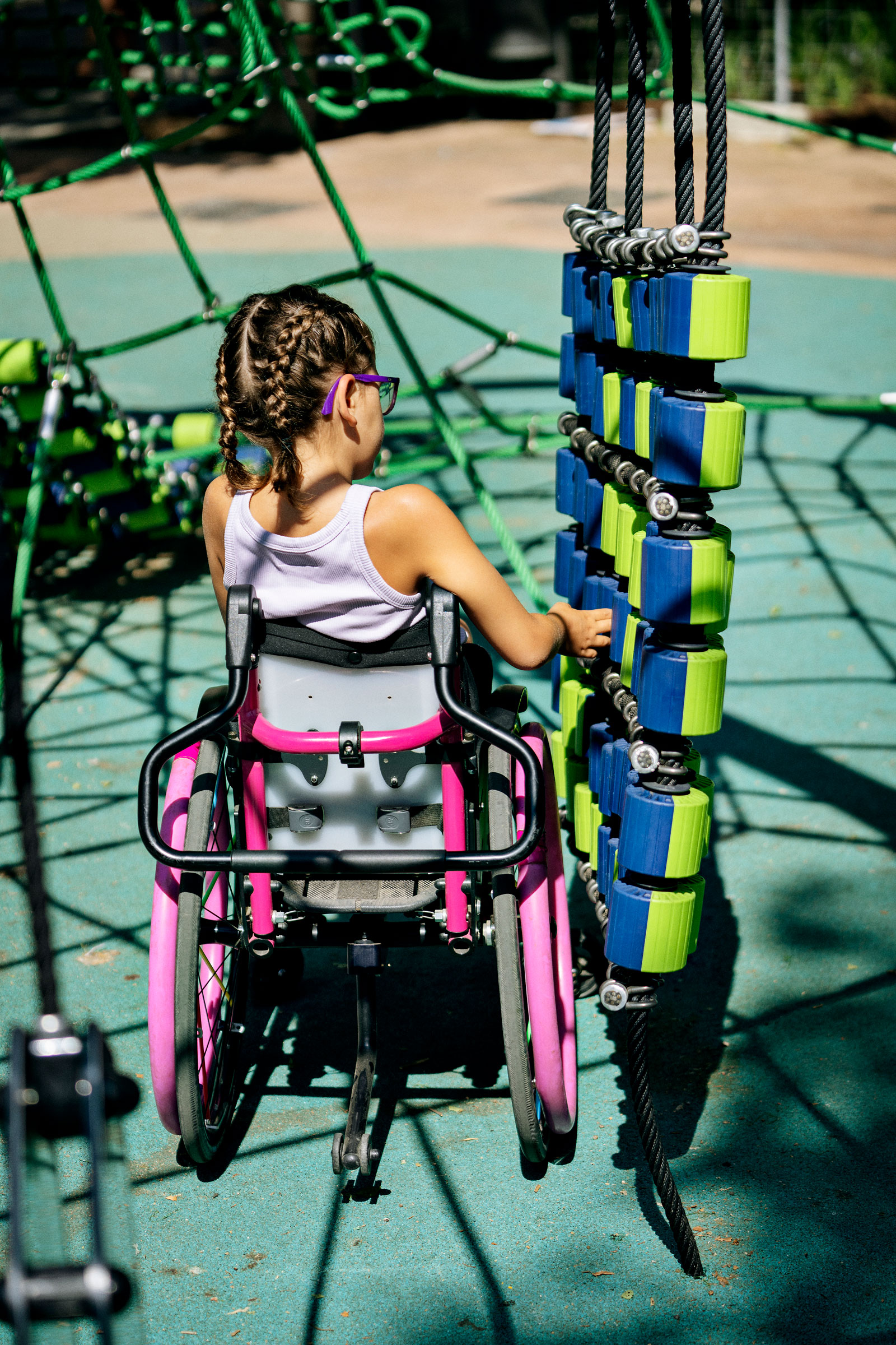 Girl in a pink wheelchair plays with blue and green climbing blocks at an inclusive outdoor playground.