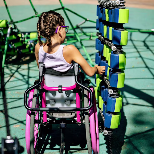 Girl in a pink wheelchair plays with blue and green climbing blocks at an inclusive outdoor playground.