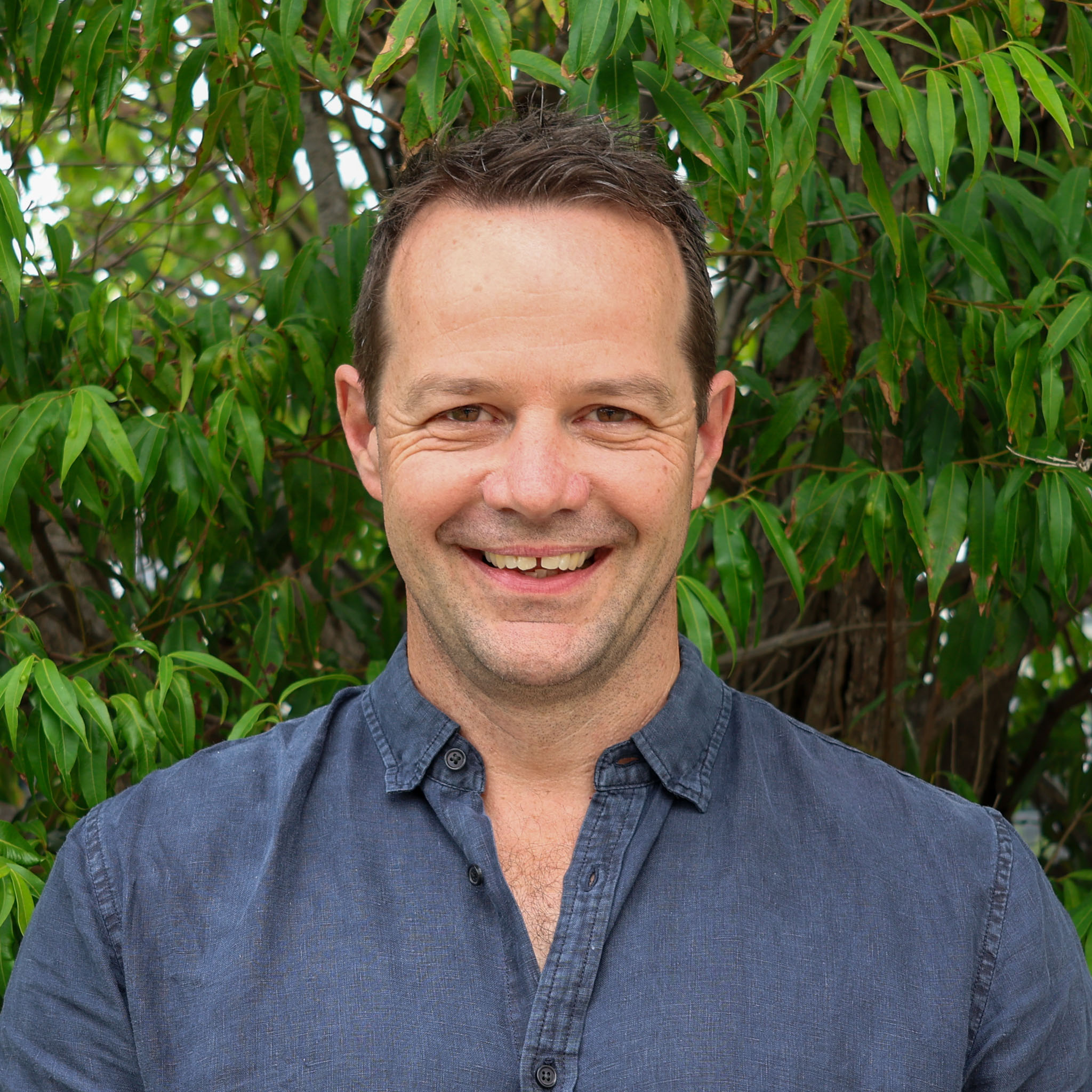 Person with blurred face wearing a blue shirt, standing in front of dense green foliage outdoors