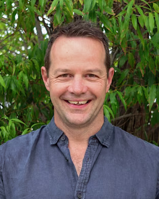 Person with blurred face wearing a blue shirt, standing in front of dense green foliage outdoors