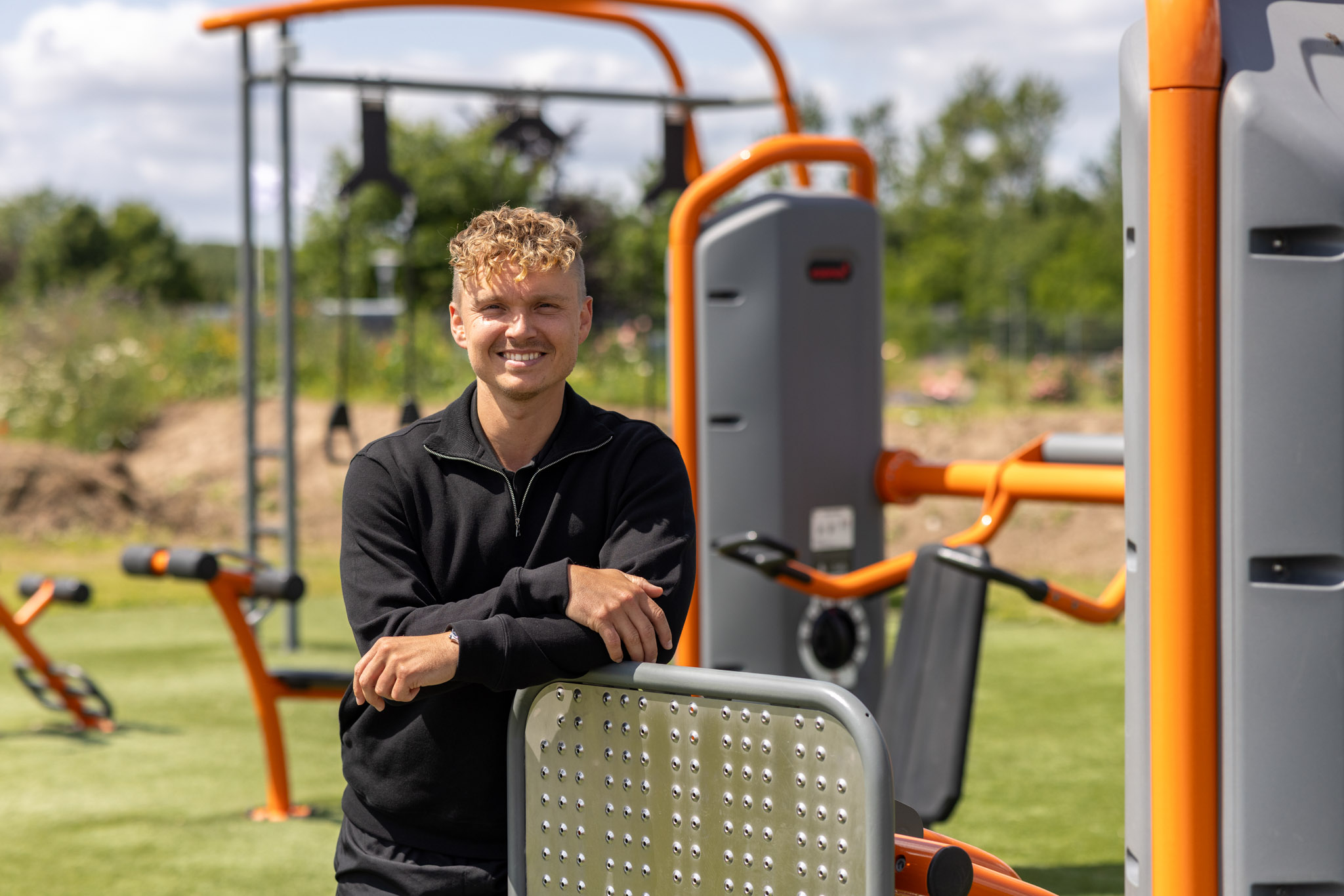 Person with blurred face in black top leaning on outdoor gym machine with orange and grey equipment on a grassy field