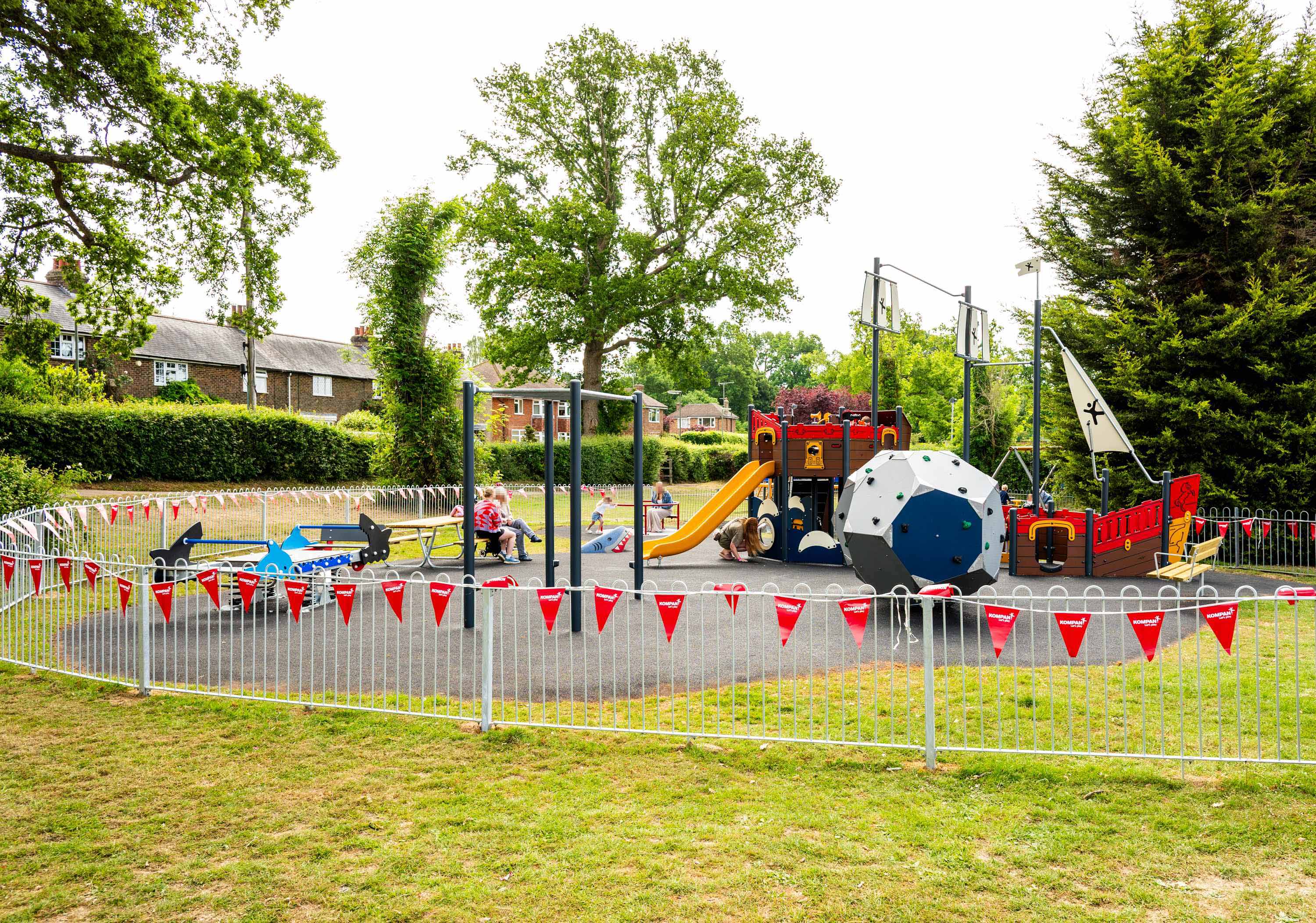 Children and parents in a fenced playground with pirate ship climbing frame, slide and colourful equipment, trees and houses behind.