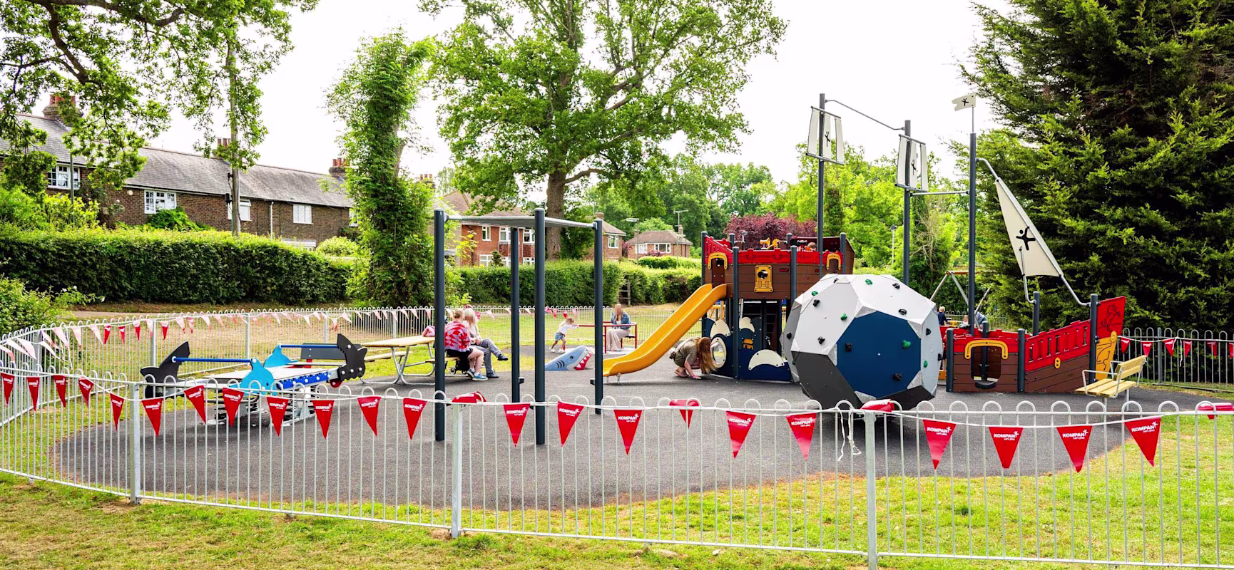 Children and parents in a fenced playground with pirate ship climbing frame, slide and colourful equipment, trees and houses behind.
