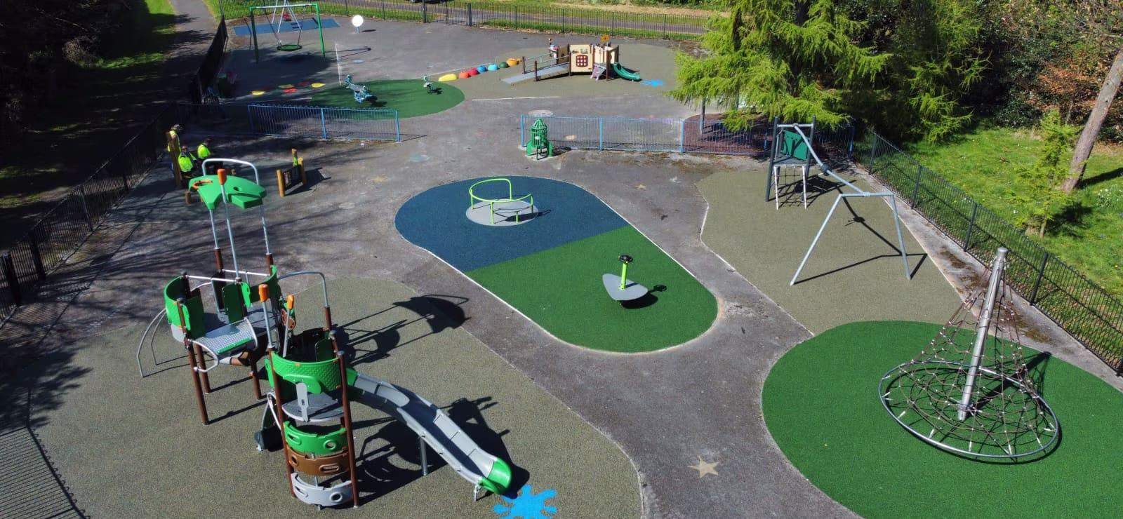 A colourful outdoor playground with slides, swings, climbing frames and roundabouts on soft surfaces, surrounded by trees and fencing.
