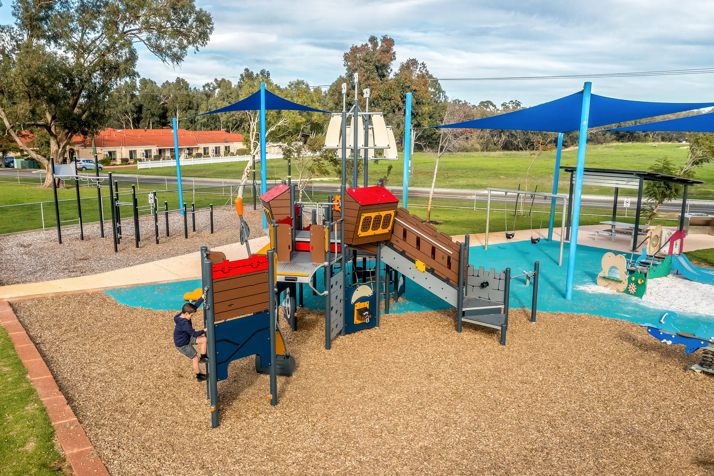 Colourful outdoor playground with climbing structures and slides under blue shade sails, with one child climbing and grassy park beyond