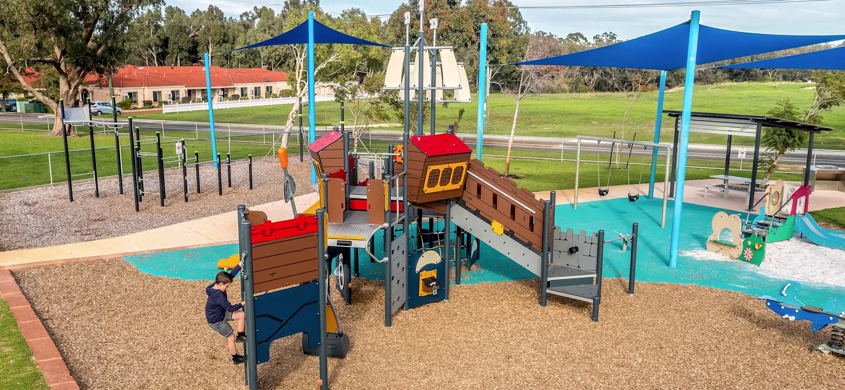 Colourful outdoor playground with climbing structures and slides under blue shade sails, with one child climbing and grassy park beyond