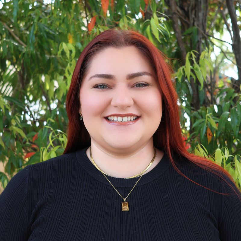 Person with long red hair in a black top and gold necklaces stands outdoors in front of green foliage and tree branches