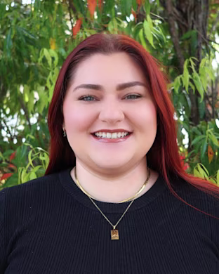 Person with long red hair in a black top and gold necklaces stands outdoors in front of green foliage and tree branches