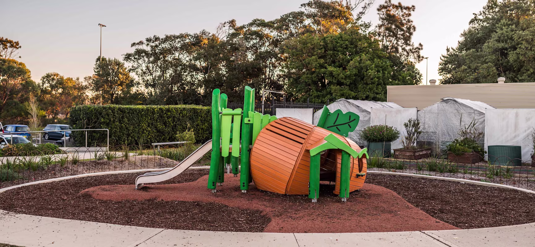 Orange and green pumpkin-shaped playground structure with slide in a park, surrounded by paths, gardens, trees, and parked cars