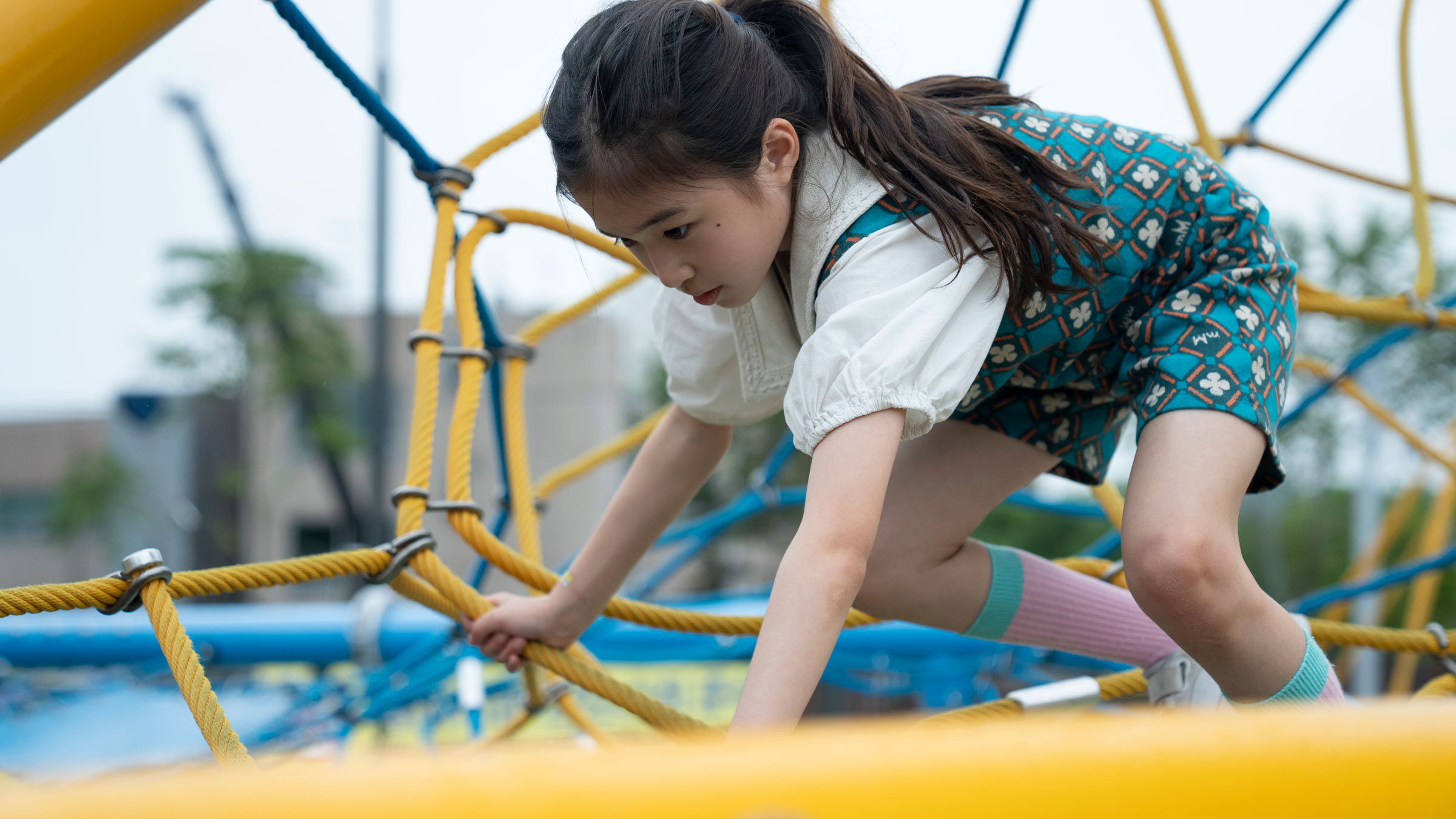 Child climbing on yellow and blue rope playground structure outdoors, wearing patterned dress and knee-high socks.