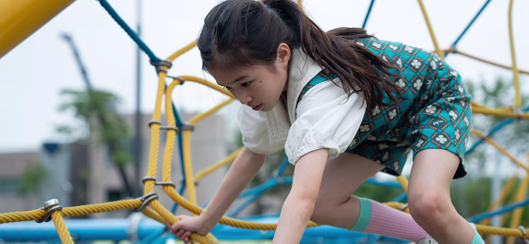 Child climbing on yellow and blue rope playground structure outdoors, wearing patterned dress and knee-high socks.