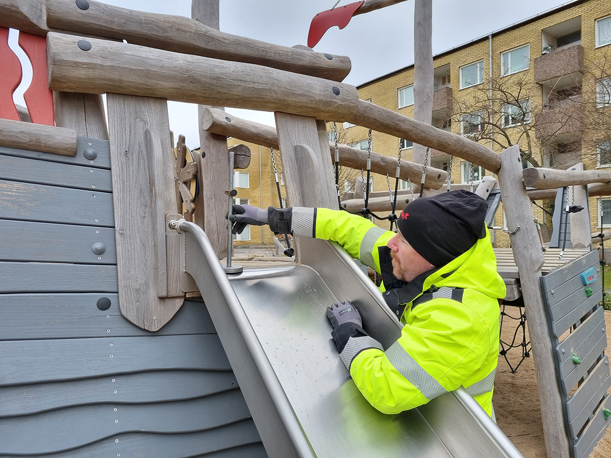 Worker in bright yellow jacket inspecting a metal slide on a wooden playground structure in an urban outdoor area.