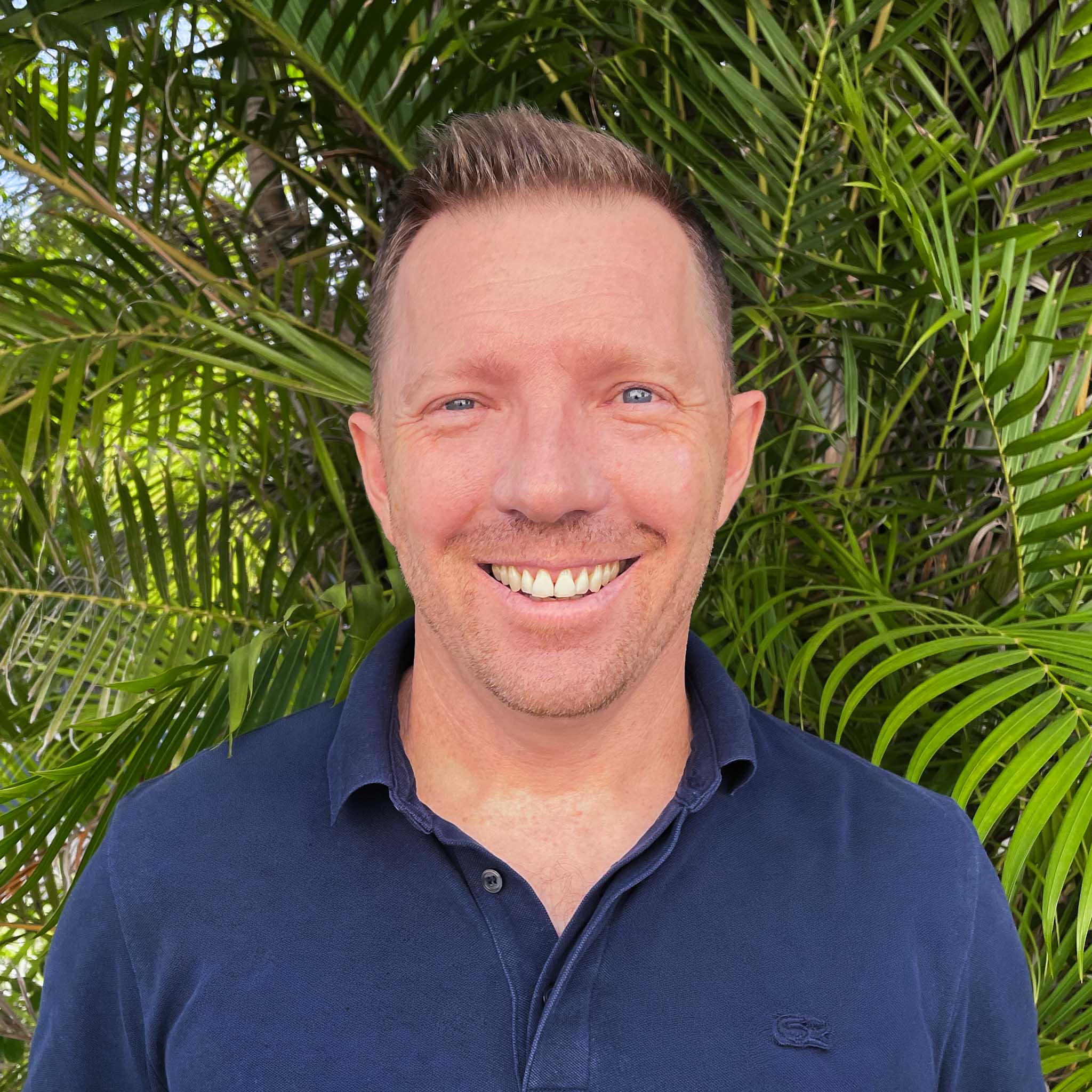 Person in navy polo shirt standing in front of dense green palm leaves, with face blurred for privacy