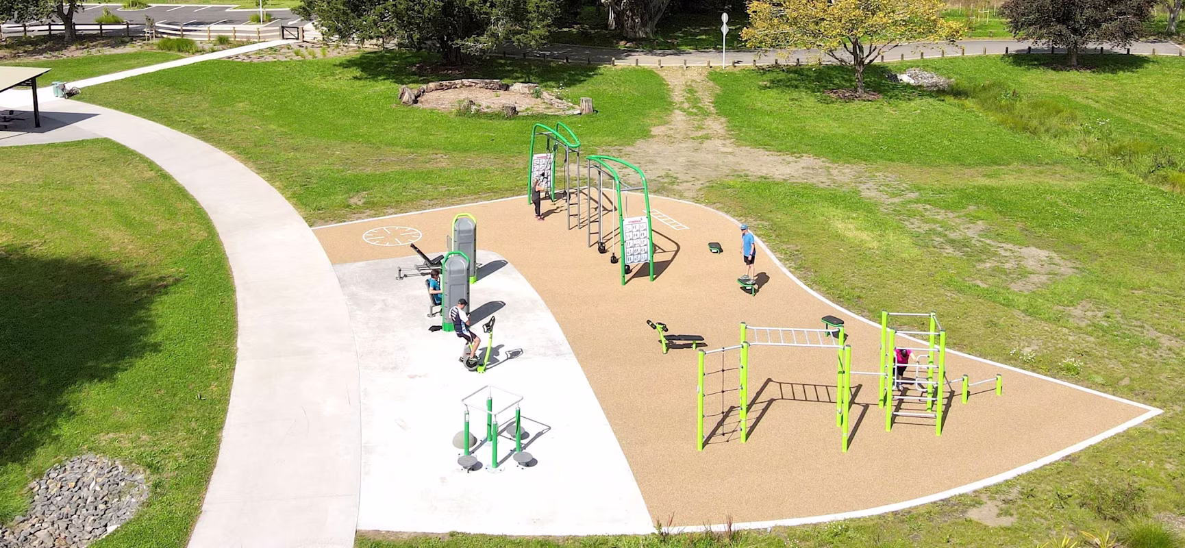 Aerial view of an outdoor green fitness park with exercise equipment, a curved path, and people working out among trees and grass