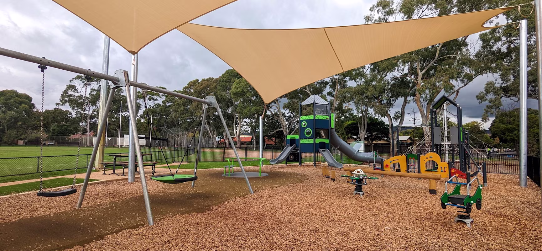 Shaded playground with swings, slides and rocking toys on woodchips, beside a green sports field and tall trees under cloudy sky