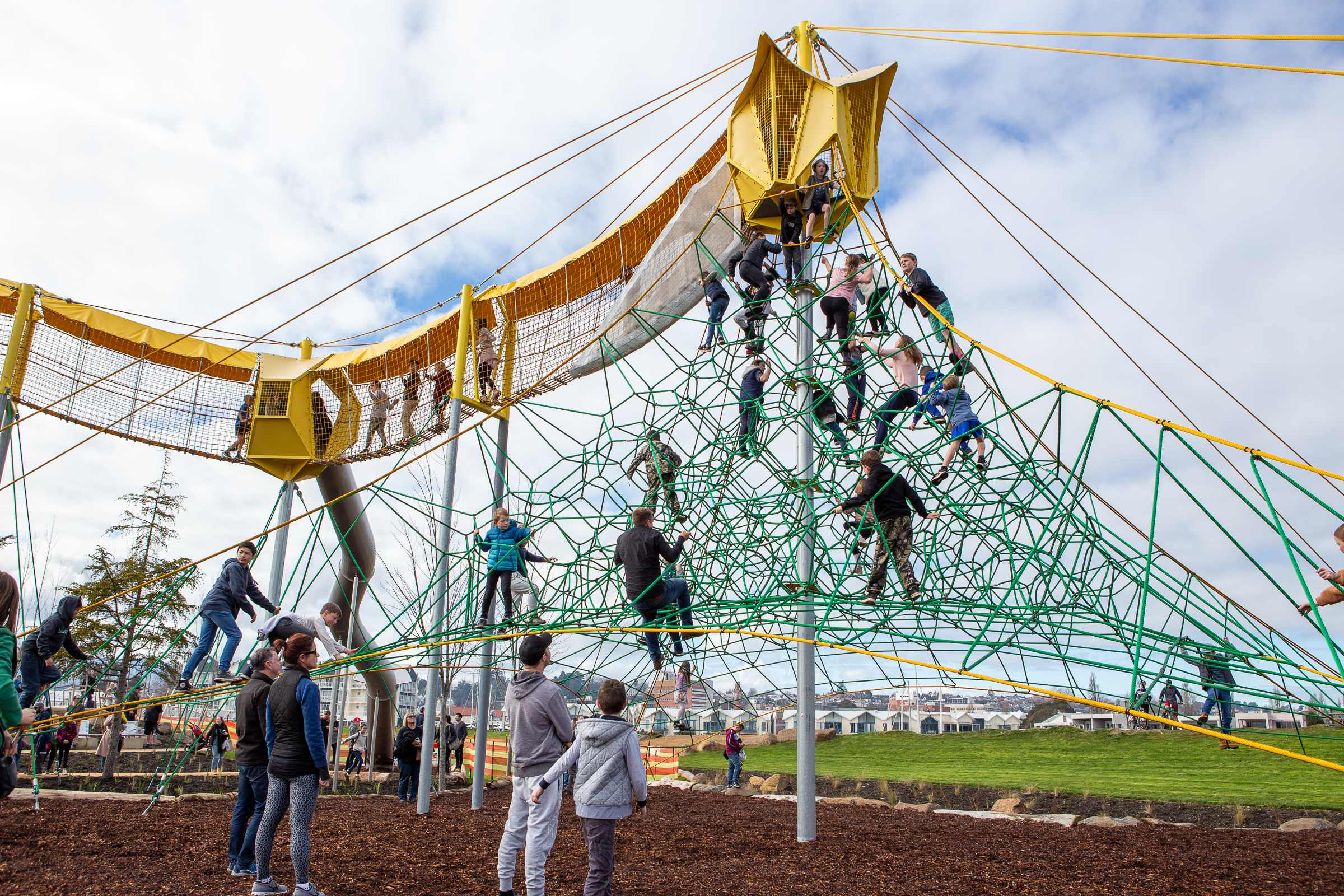 Children climb a large green rope pyramid and walk a yellow elevated net bridge at a busy outdoor playground under a cloudy sky.