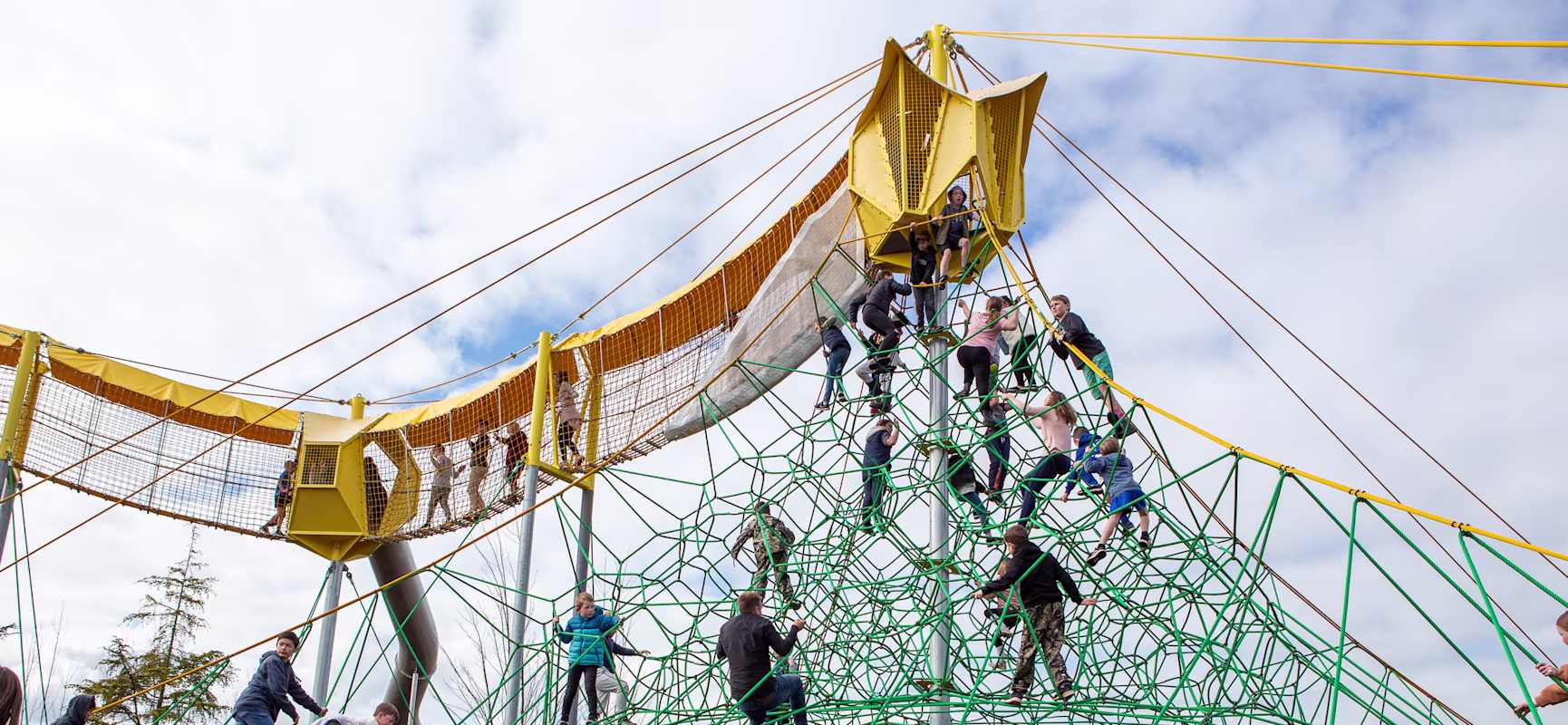 Children climb a large green rope pyramid and walk a yellow elevated net bridge at a busy outdoor playground under a cloudy sky.