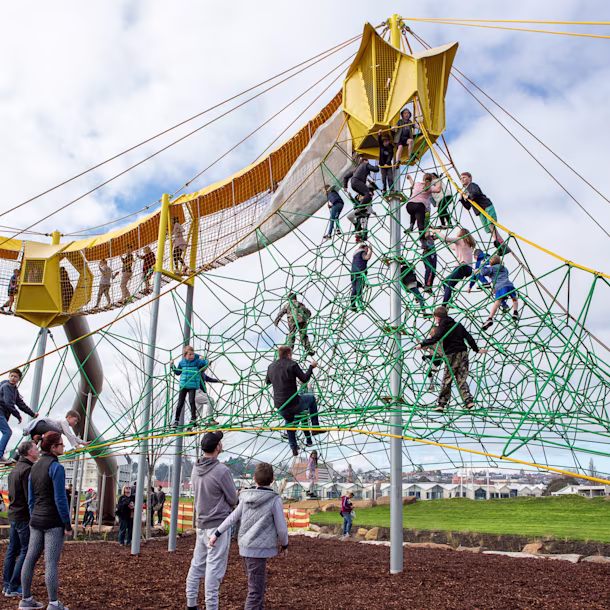 Children climb a large green rope pyramid and walk a yellow elevated net bridge at a busy outdoor playground under a cloudy sky.