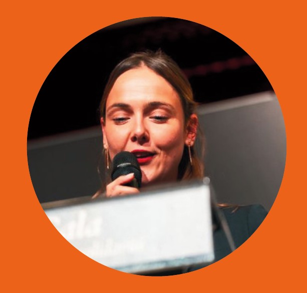 Person with blurred face speaking at a podium against a dark background, framed in a circular crop on an orange background.