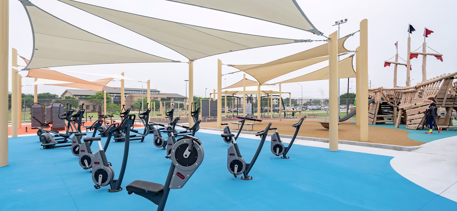 Outdoor gym with stationary bikes on bright blue floor under shade sails, beside a wooden playground and open park area