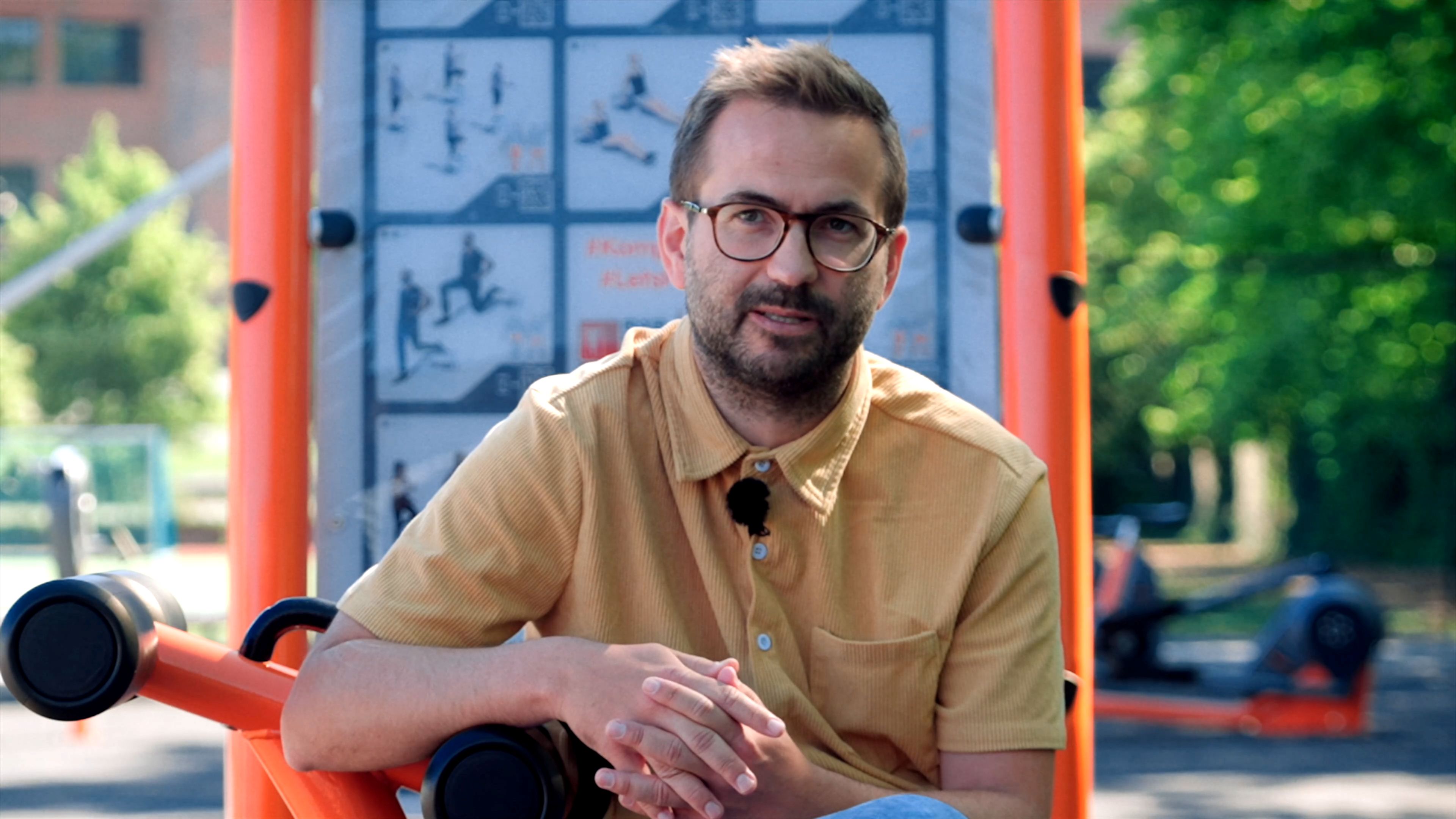Person with blurred face sitting on outdoor gym equipment, wearing a tan shirt, with exercise instruction board and trees in background.